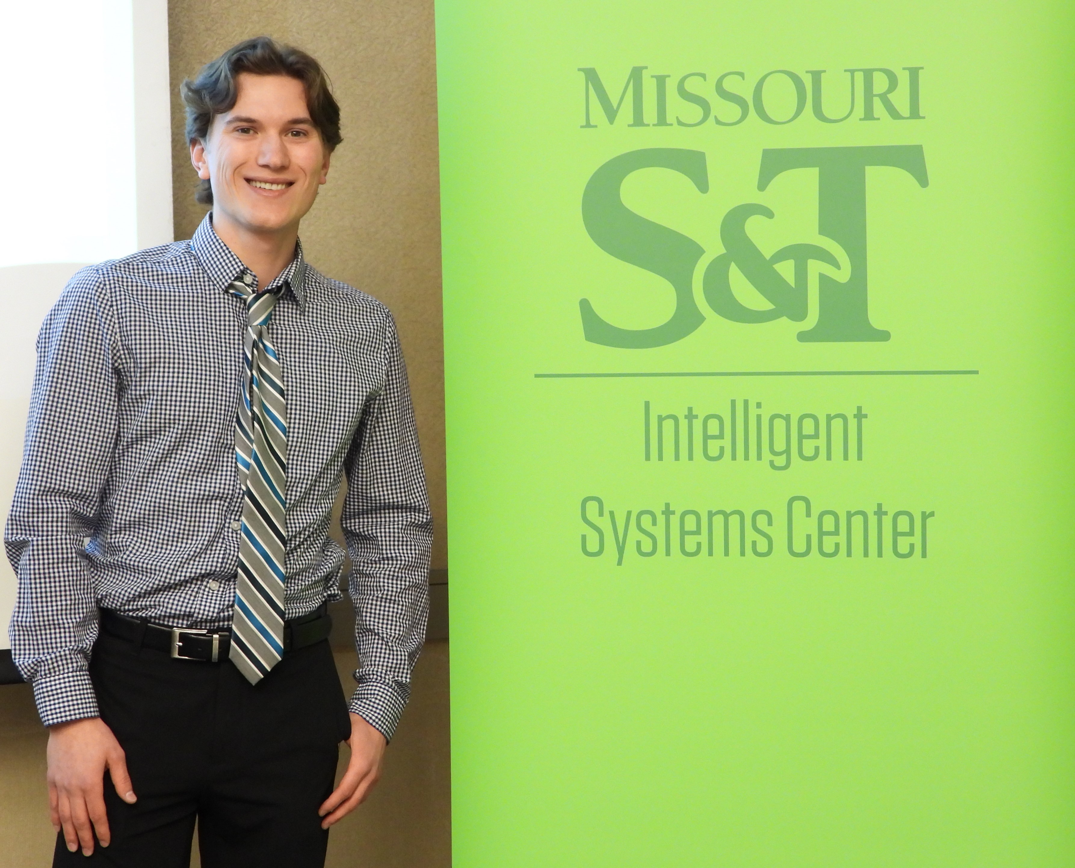 Graduate researcher, Colton Walker, smiling in front of Intelligent Systems Center banner.